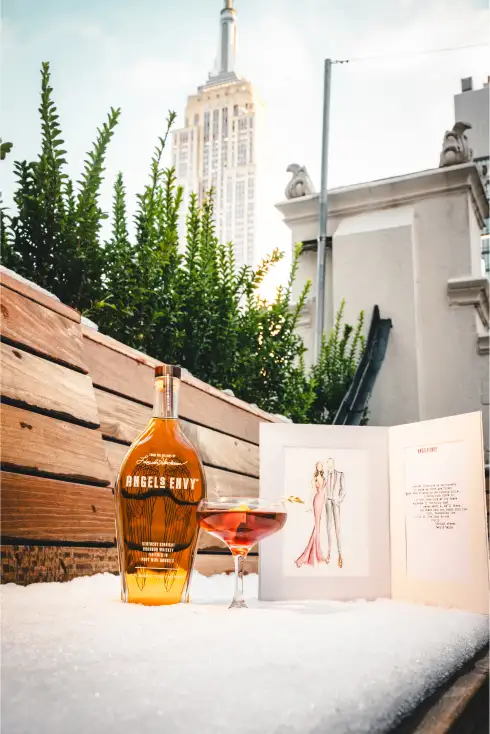 A bottle of Angel’s Envy, a cocktail, and a custom greeting card on a rooftop in NYC, with the Empire State Building in the background
