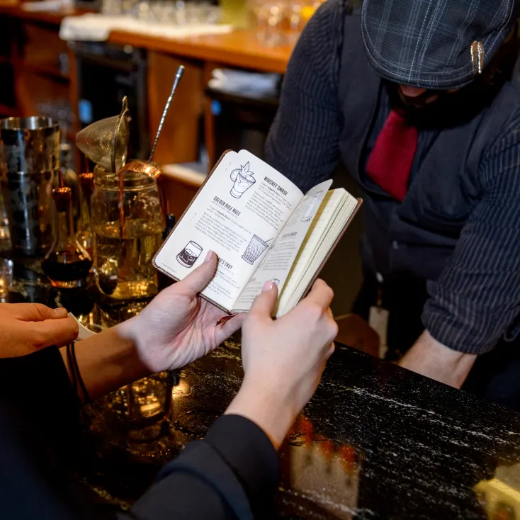 A person holding a recipe book in front of a bartender at a bar