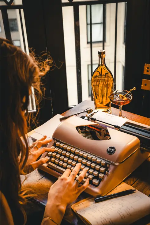 An individual typing at a typewriter with a bottle of Angel’s Envy in the background
